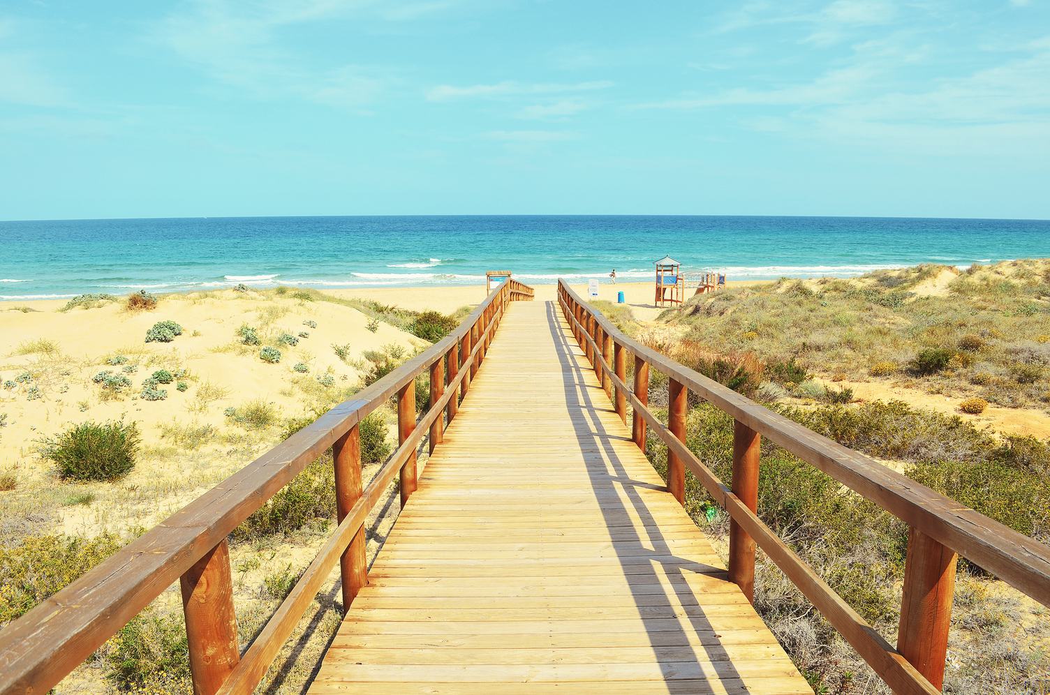 Elevated timber beach walkover with straight railings crossing open sandy dunes toward turquoise ocean under blue sky.